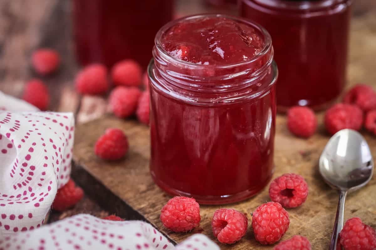 Pot of Seedless Raspberry Jam surrounded by raspberries on a wooden board next to a teaspoon.