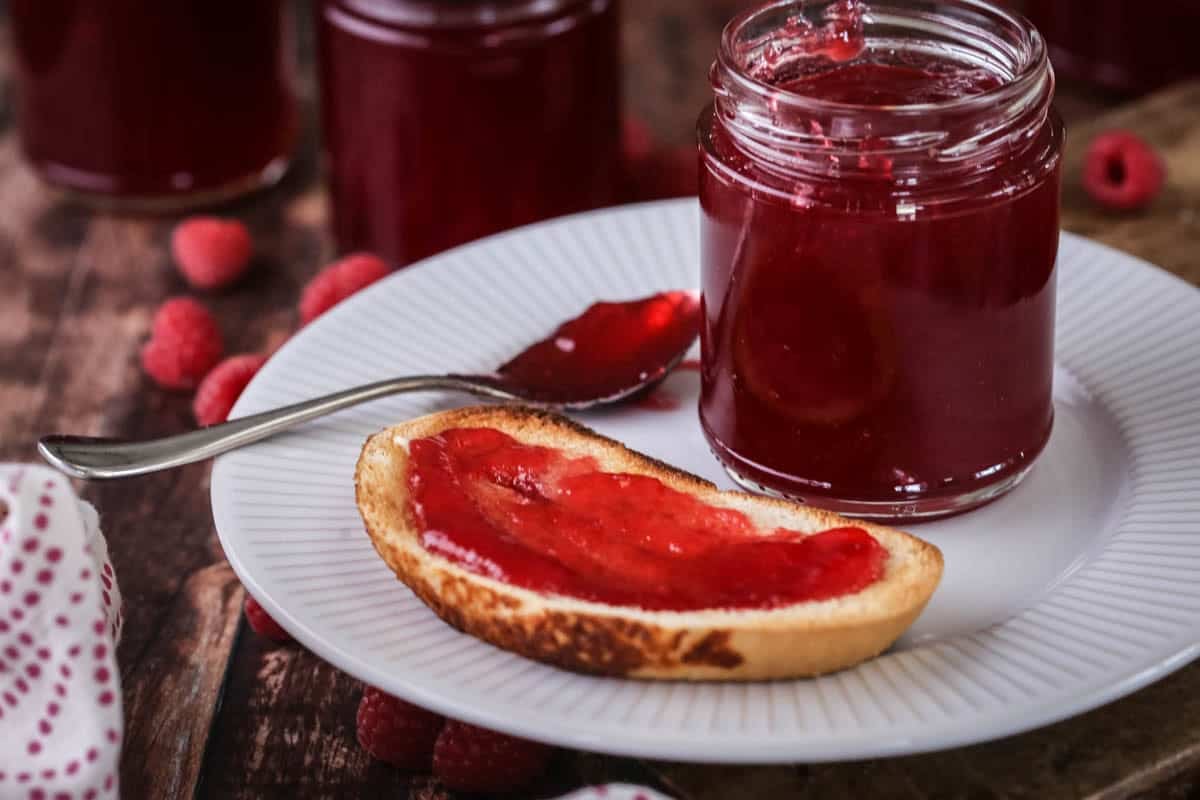 Jam spread on a slice of toast showing bright vibrant raspberry jam spread with no seeds. On a plate with a pot of jam and teaspoon.