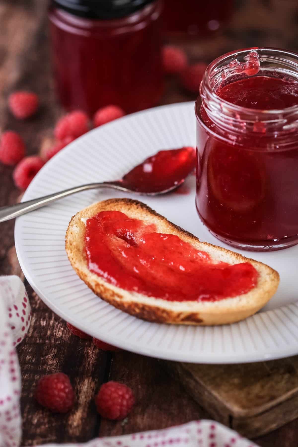 Jam spread on a slice of toast showing bright vibrant raspberry jam spread with no seeds. On a plate with a pot of jam and teaspoon.