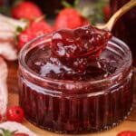 Spoon scooping up strawberry and redcurrant jam from a glass pot.