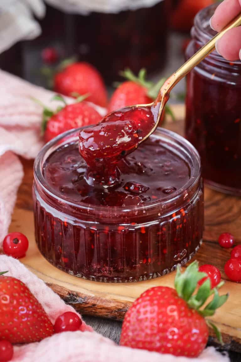 Spoon scooping up strawberry and redcurrant jam from a glass pot.
