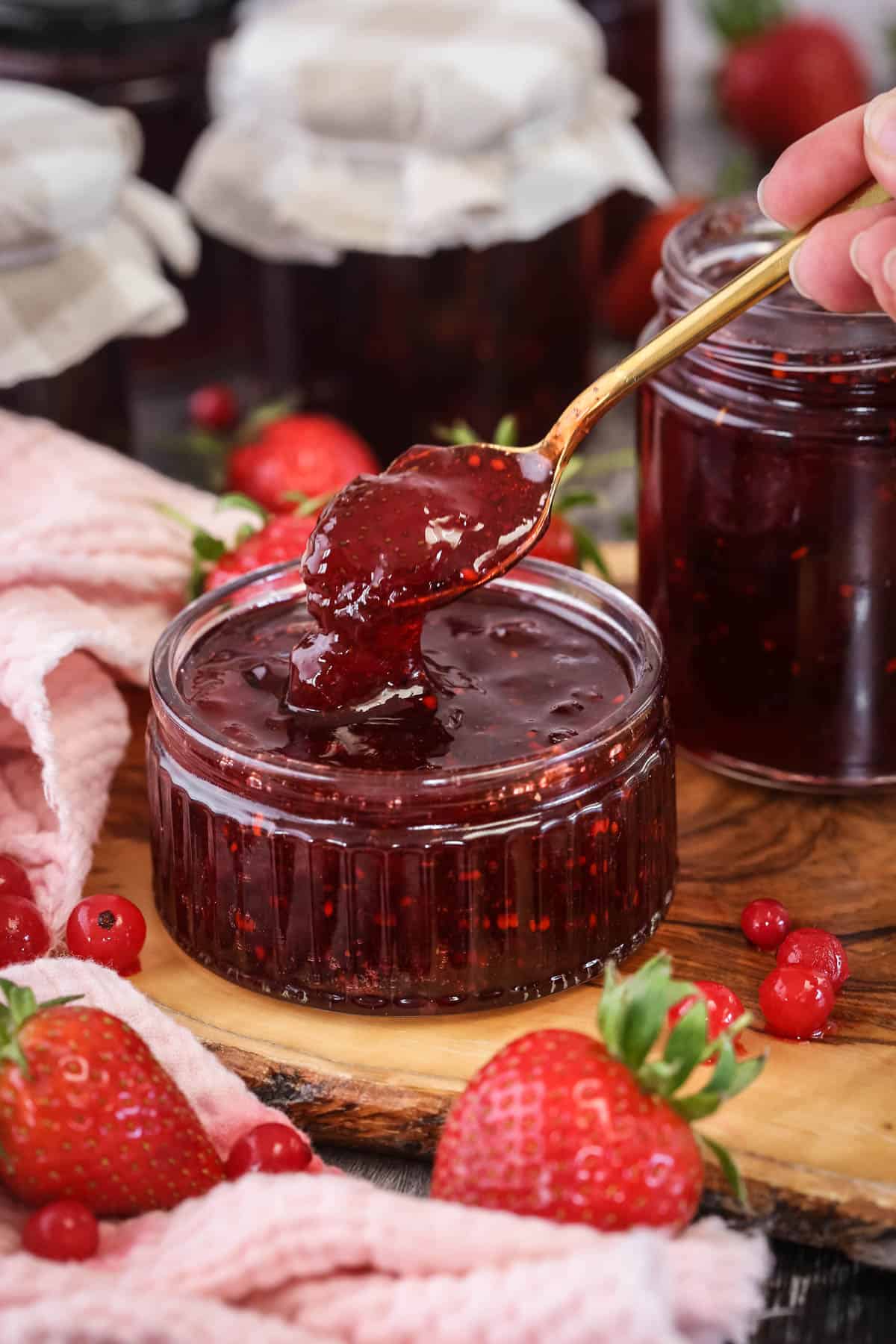 Spoon scooping up strawberry and redcurrant jam from a glass pot.