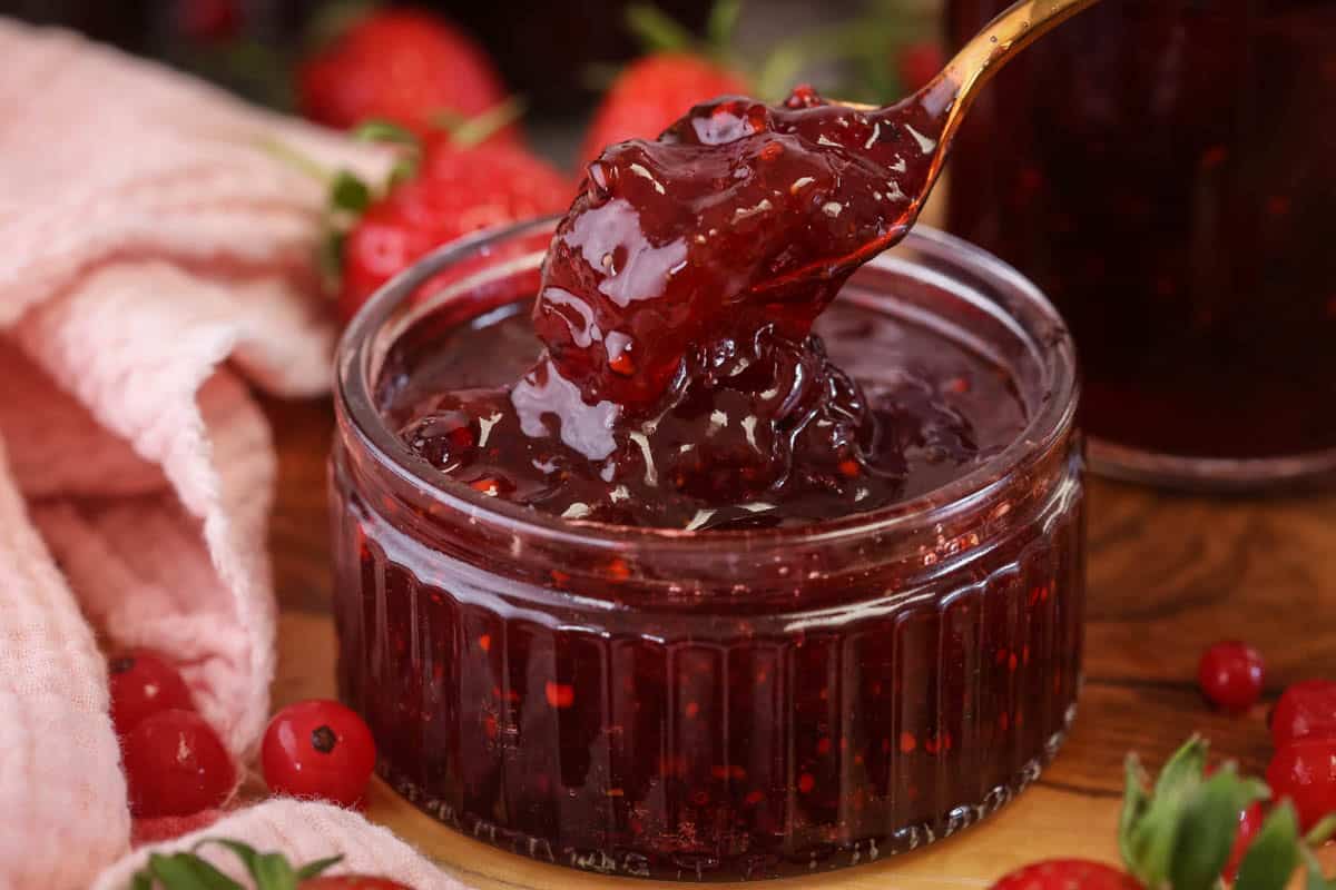 Spoon scooping up strawberry and redcurrant jam from a glass pot.