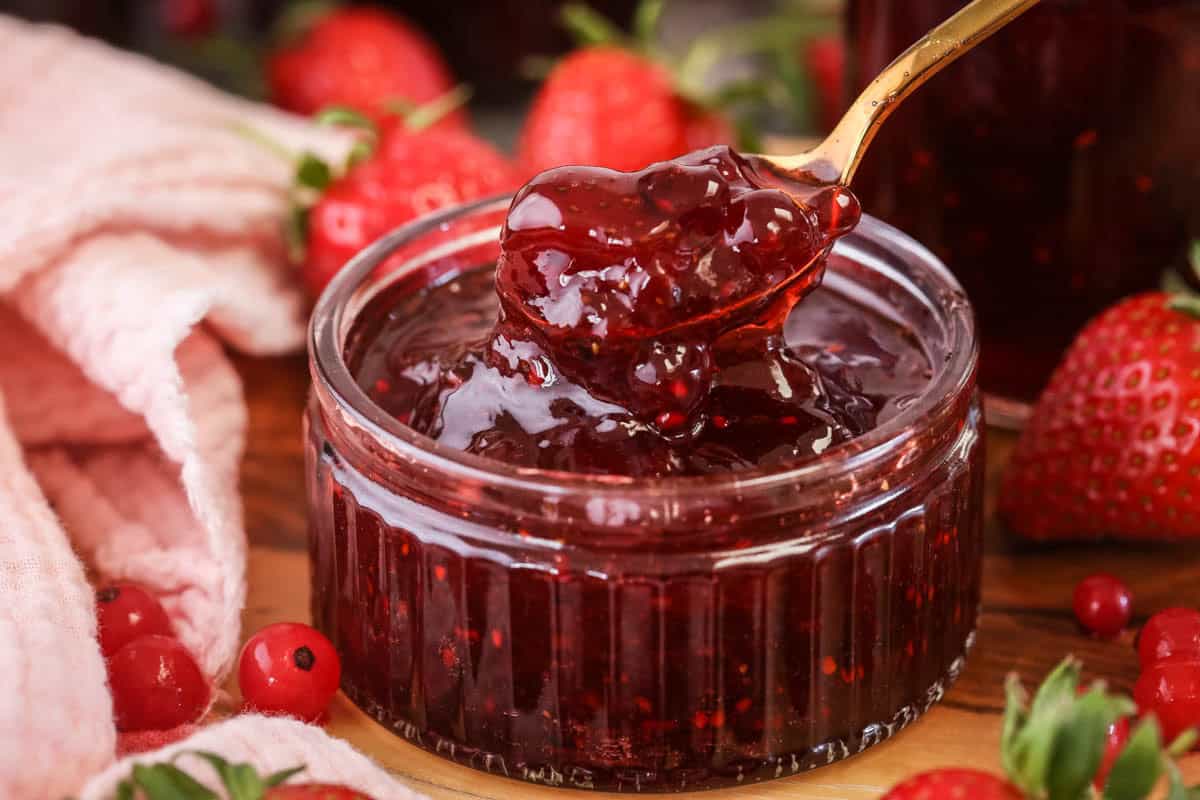 Spoon scooping up strawberry and redcurrant jam from a glass pot.