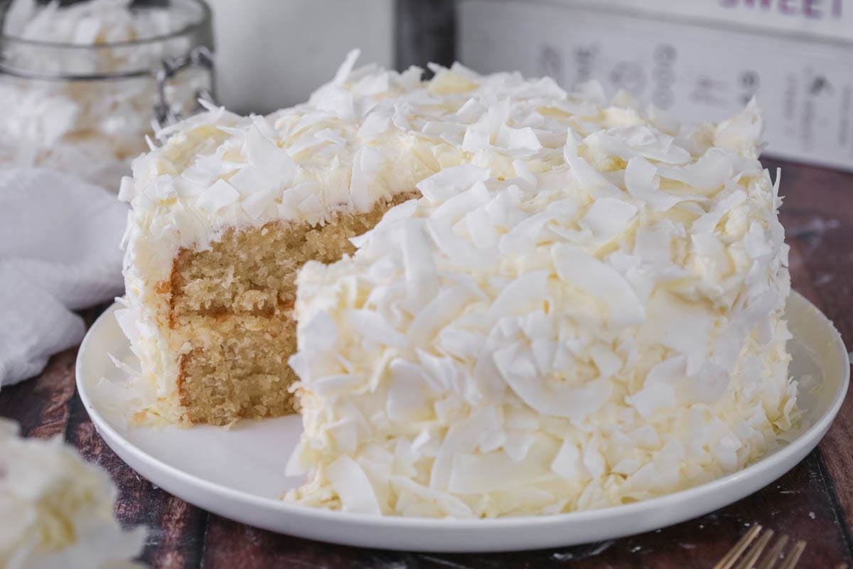 Gluten-Free Coconut Cake covered in coconut flakes on a white plate. Cut into showing soft moisture texture.