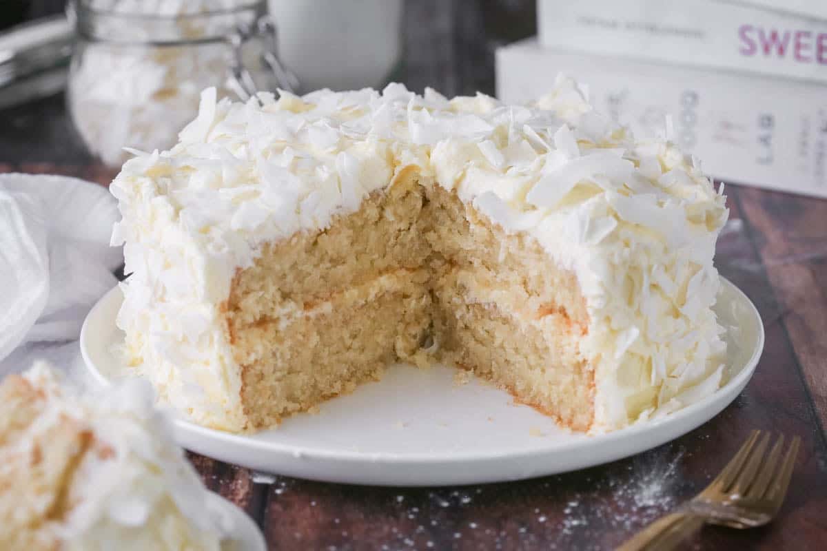 Gluten-Free Coconut Cake covered in coconut flakes on a white plate. Cut into showing soft moisture texture.