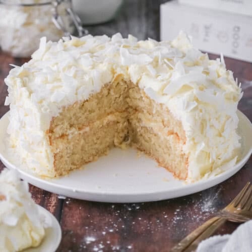 Gluten-Free Coconut Cake covered in coconut flakes on a white plate. Cut into showing soft moisture texture.
