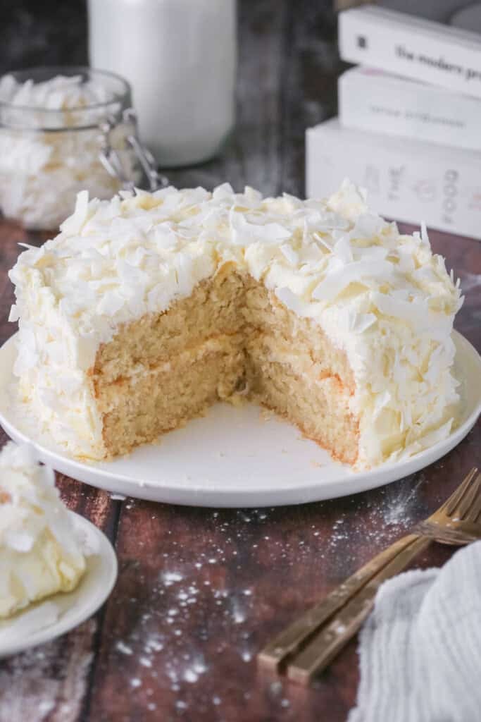 Gluten-Free Coconut Cake covered in coconut flakes on a white plate. Cut into showing soft moisture texture.