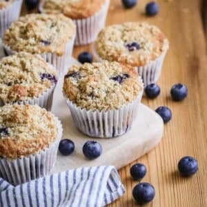 Freshly baked Blueberry Muffins with golden streusel topping on a wooden board on a table surrounded by blueberries.