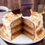 Gluten Free S'mores Cake cut to reveal three layers of cinnamon sponge, filled with plant-based chocolate buttercream and covered with toasted marshmallow meringue. Decorated with chocolate shards and gluten-free biscuit crumbs.