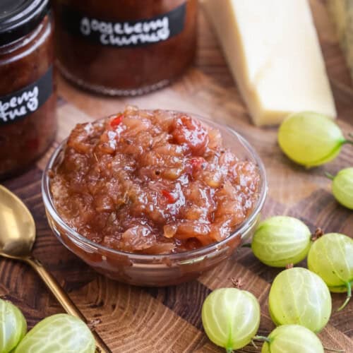 Gooseberry Chutney in a small dish, surrounded by fresh gooseberries, cheese and jars of chutney.