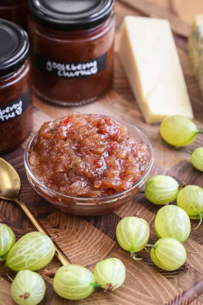 Gooseberry Chutney in a small dish, surrounded by fresh gooseberries, cheese and jars of chutney.