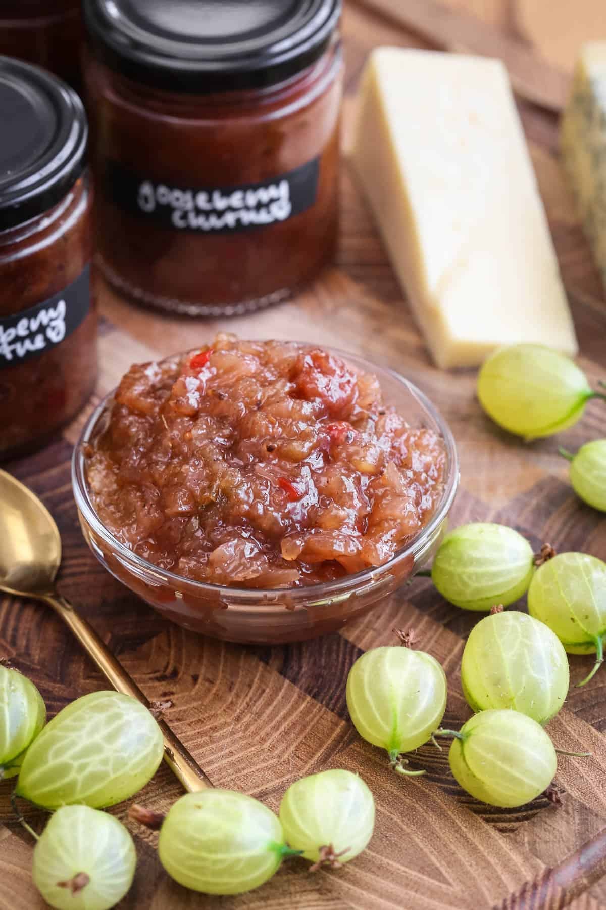 Gooseberry Chutney in a small dish, surrounded by fresh gooseberries, cheese and jars of chutney.