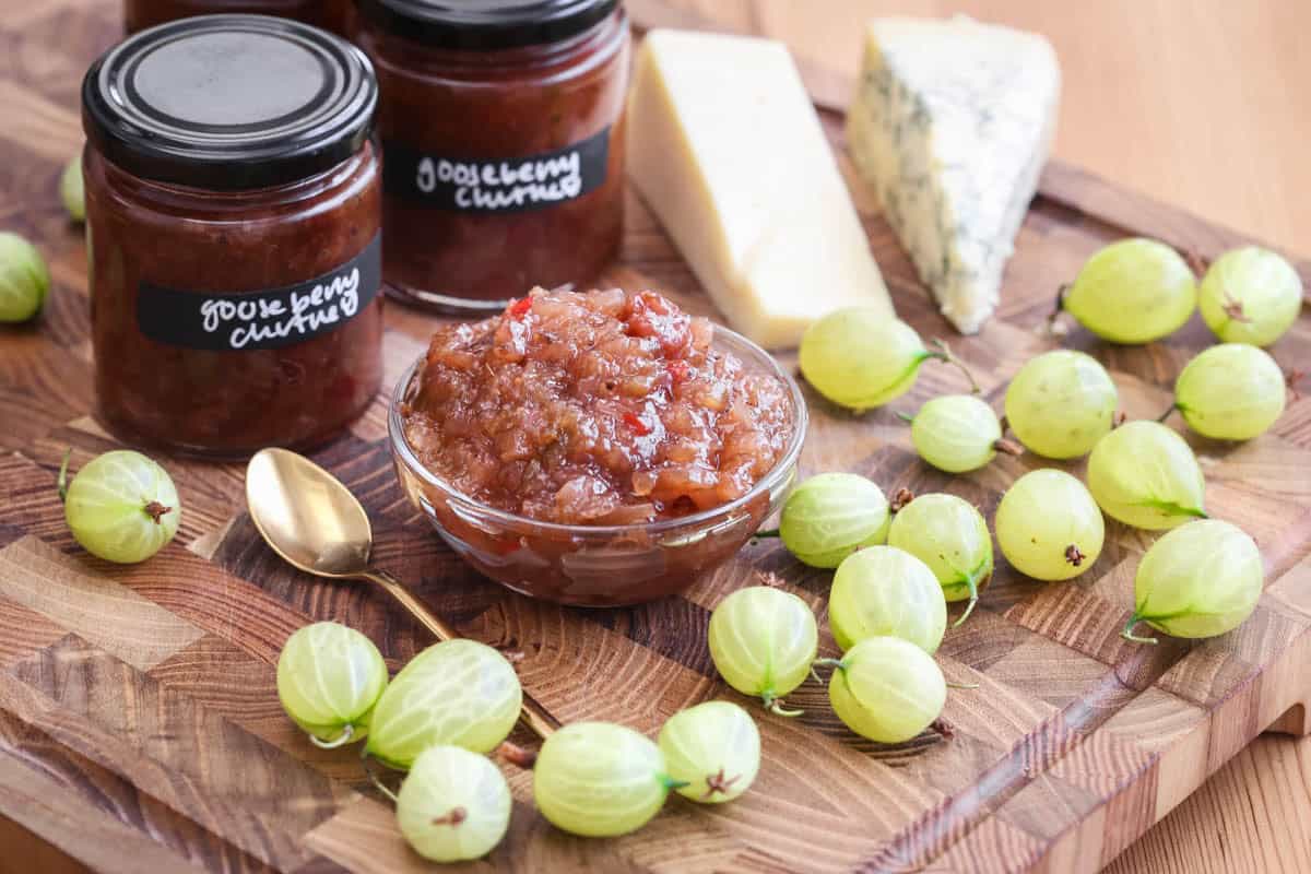 Gooseberry Chutney in a small dish, surrounded by fresh gooseberries, cheese and jars of chutney.