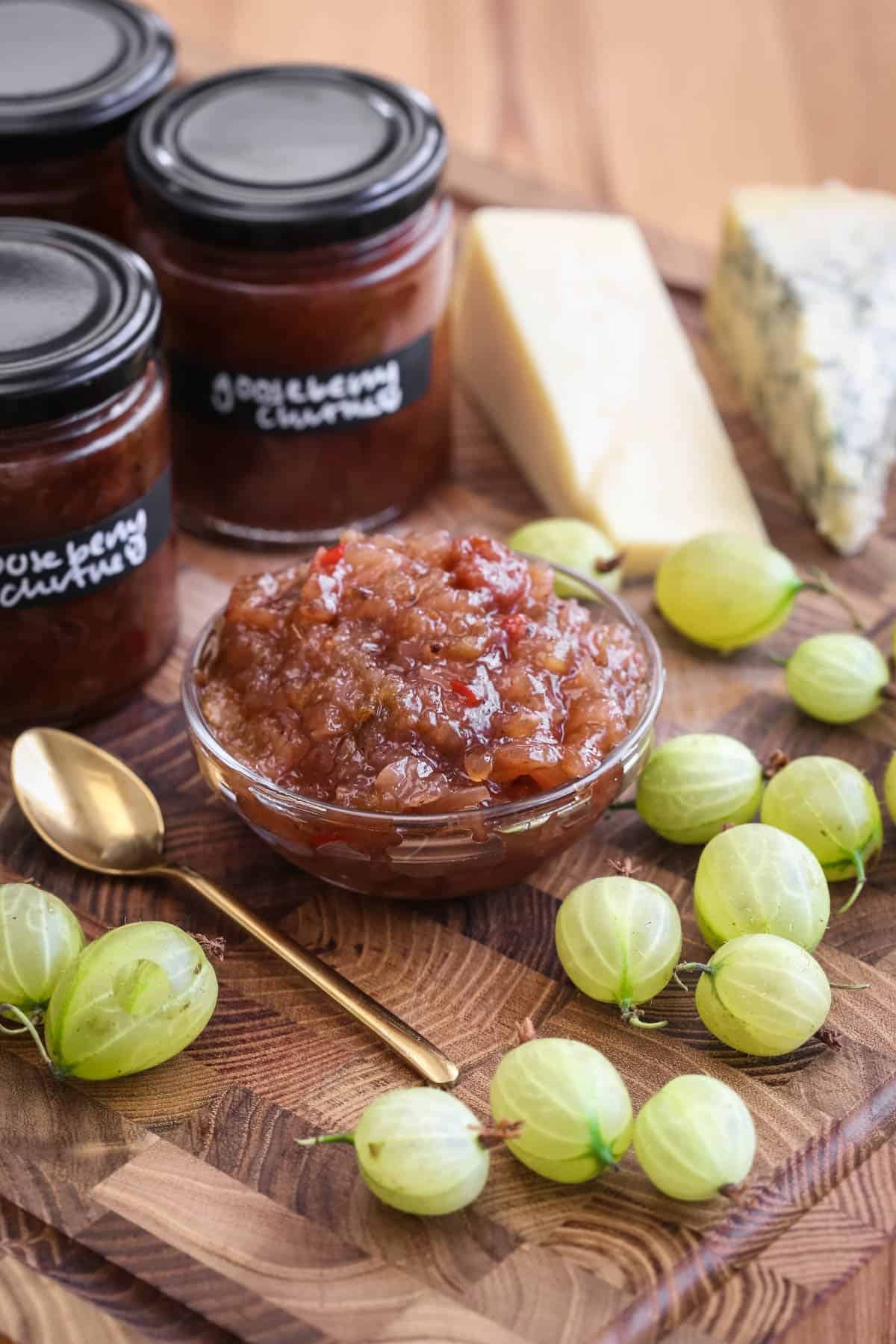 Gooseberry Chutney in a small dish, surrounded by fresh gooseberries, cheese and jars of chutney.