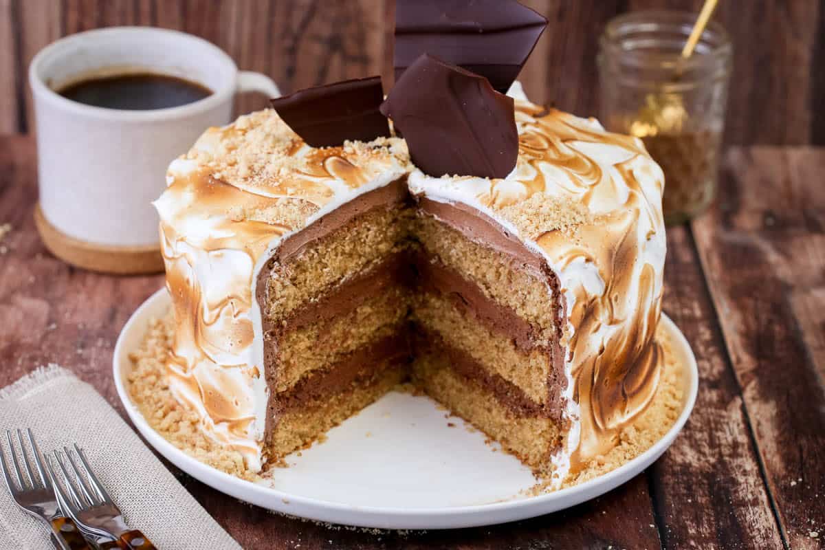 Gluten Free S'mores Cake cut to reveal three layers of cinnamon sponge, filled with plant-based chocolate buttercream and covered with toasted marshmallow meringue. Decorated with chocolate shards and gluten-free biscuit crumbs.