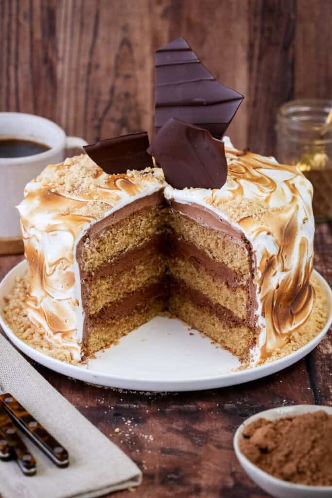Gluten Free S'mores Cake cut to reveal three layers of cinnamon sponge, filled with plant-based chocolate buttercream and covered with toasted marshmallow meringue. Decorated with chocolate shards and gluten-free biscuit crumbs.