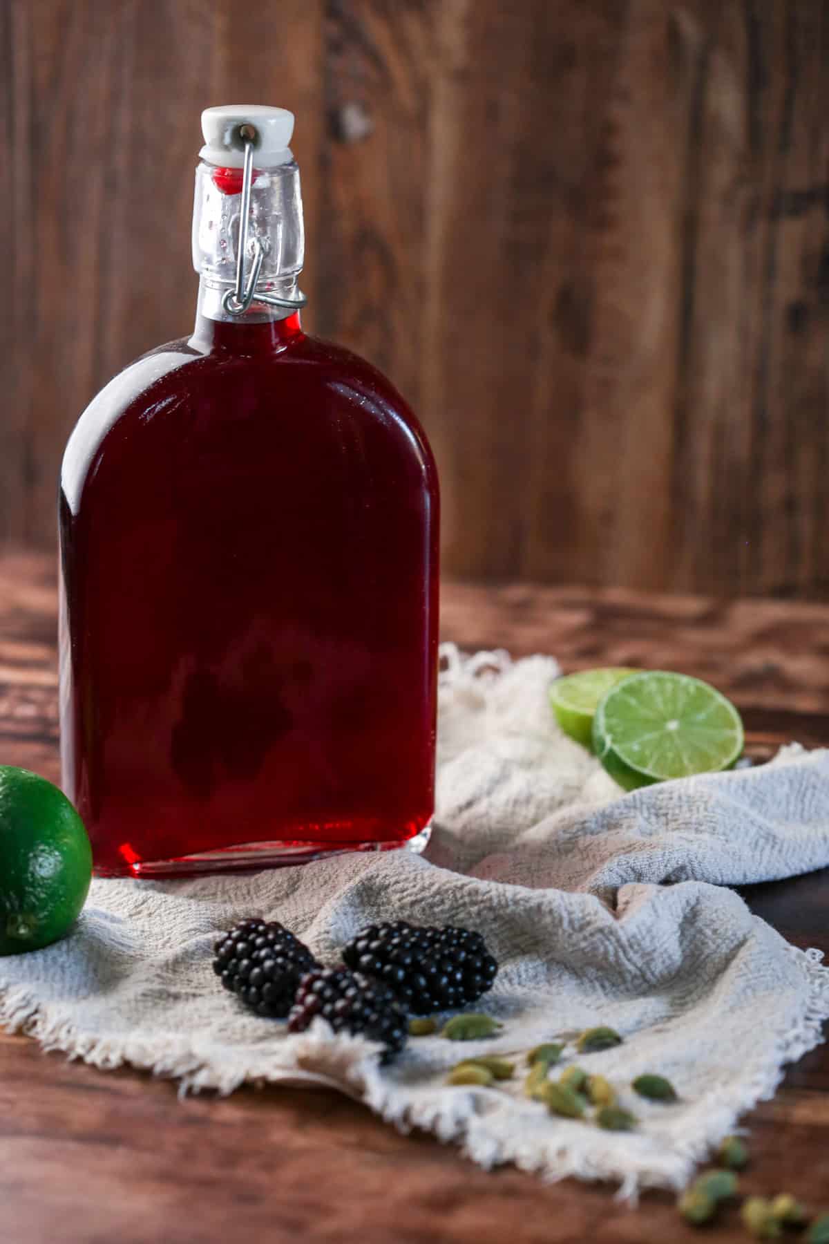 Full bottle of Blackberry Gin Liqueur surrounded by blackberries, cardamom and limes.