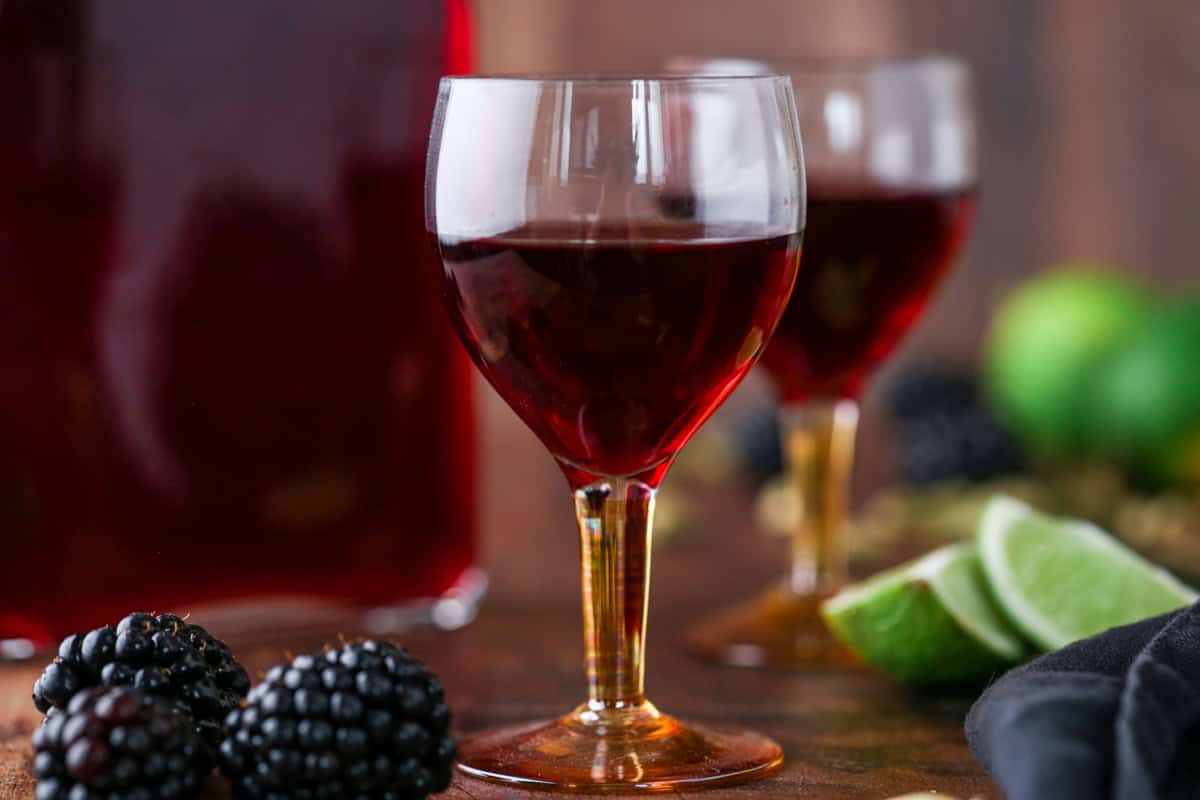 Glass of Blackberry Gin Liqueur surrounded by the full bottle, blackberries, cardamom and limes.