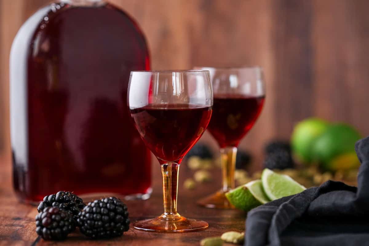 Glass of Blackberry Gin Liqueur surrounded by the full bottle, blackberries, cardamom and limes.