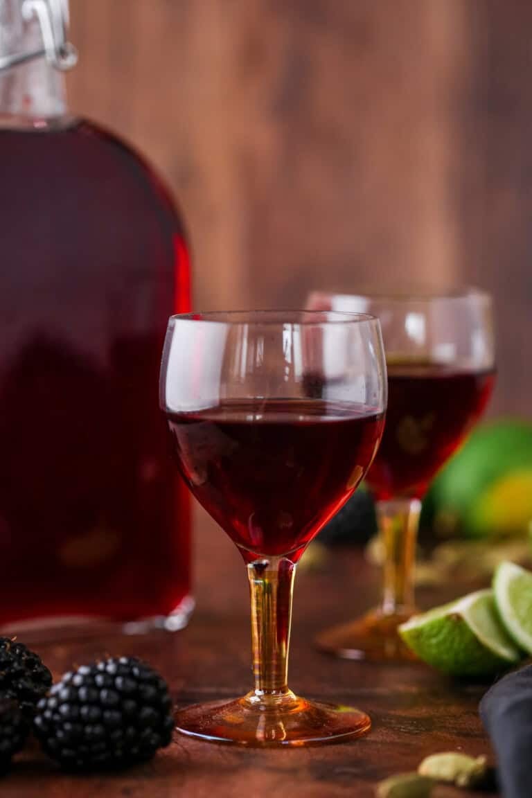 Glass of Blackberry Gin Liqueur surrounded by the full bottle, blackberries, cardamom and limes.
