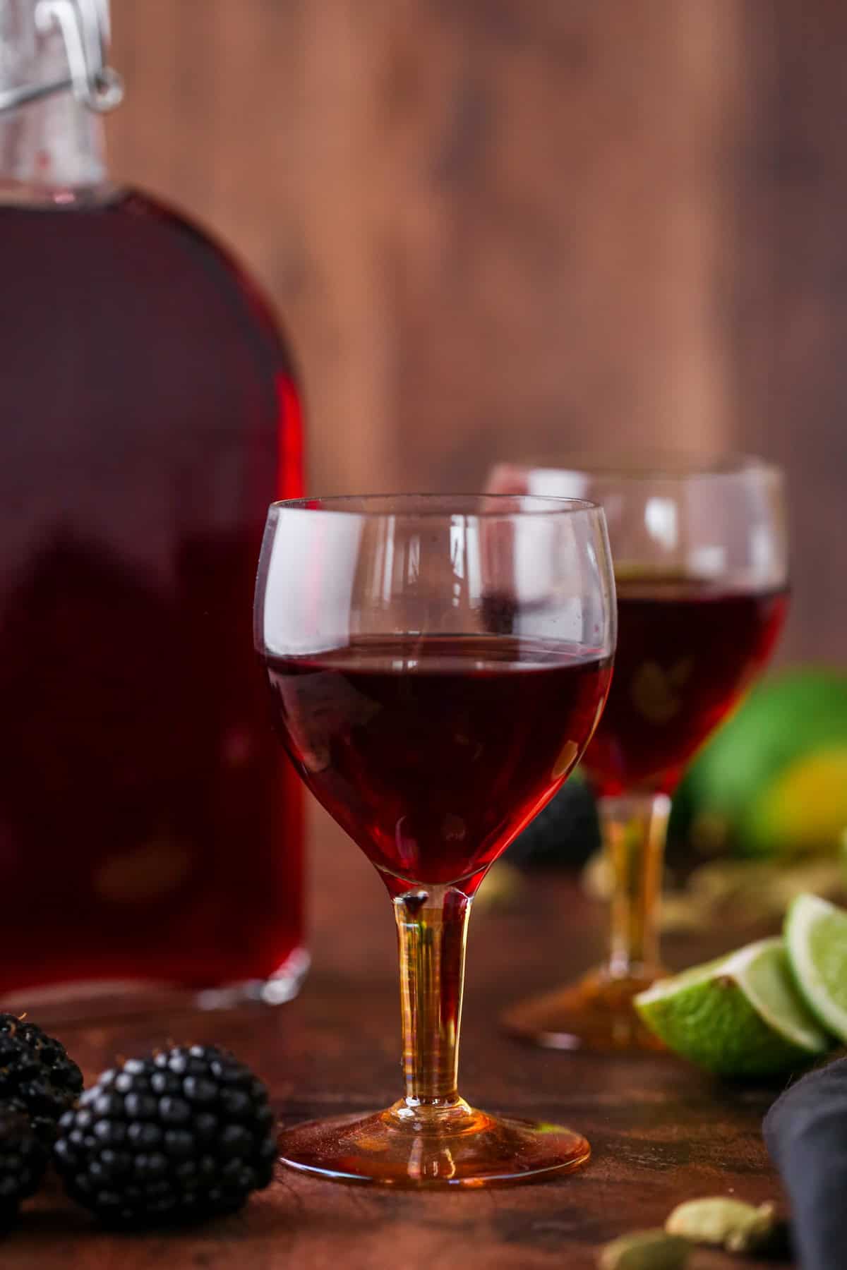 Glass of Blackberry Gin Liqueur surrounded by the full bottle, blackberries, cardamom and limes.