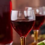 Glass of Blackberry Gin Liqueur surrounded by the full bottle, blackberries, cardamom and limes. Pin image.
