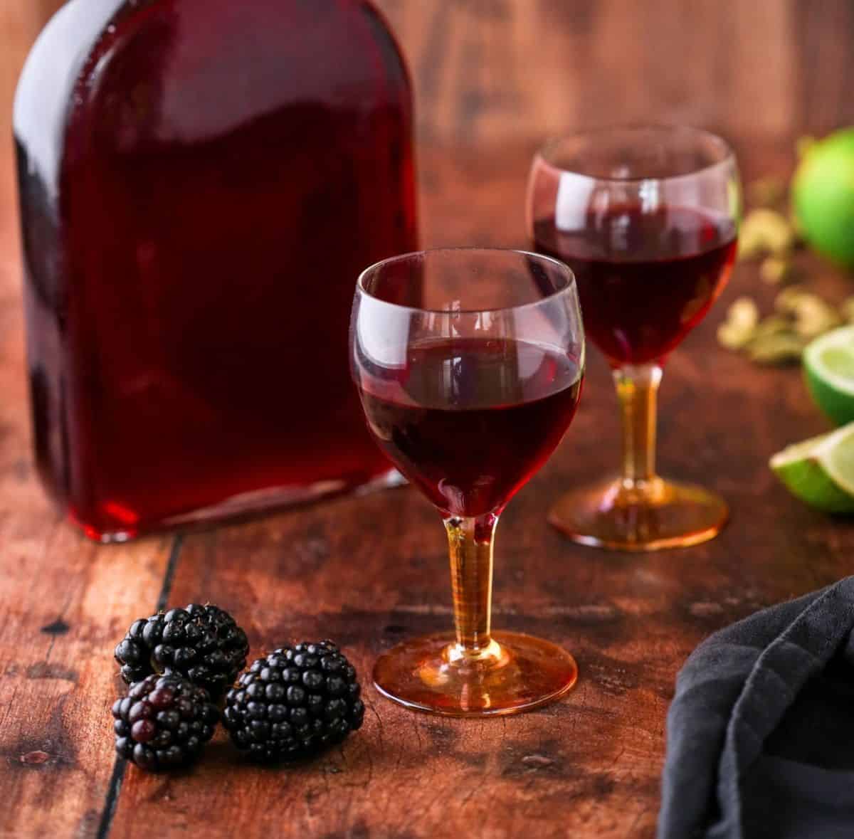 Glass of Blackberry Gin Liqueur surrounded by the full bottle, blackberries, cardamom and limes.