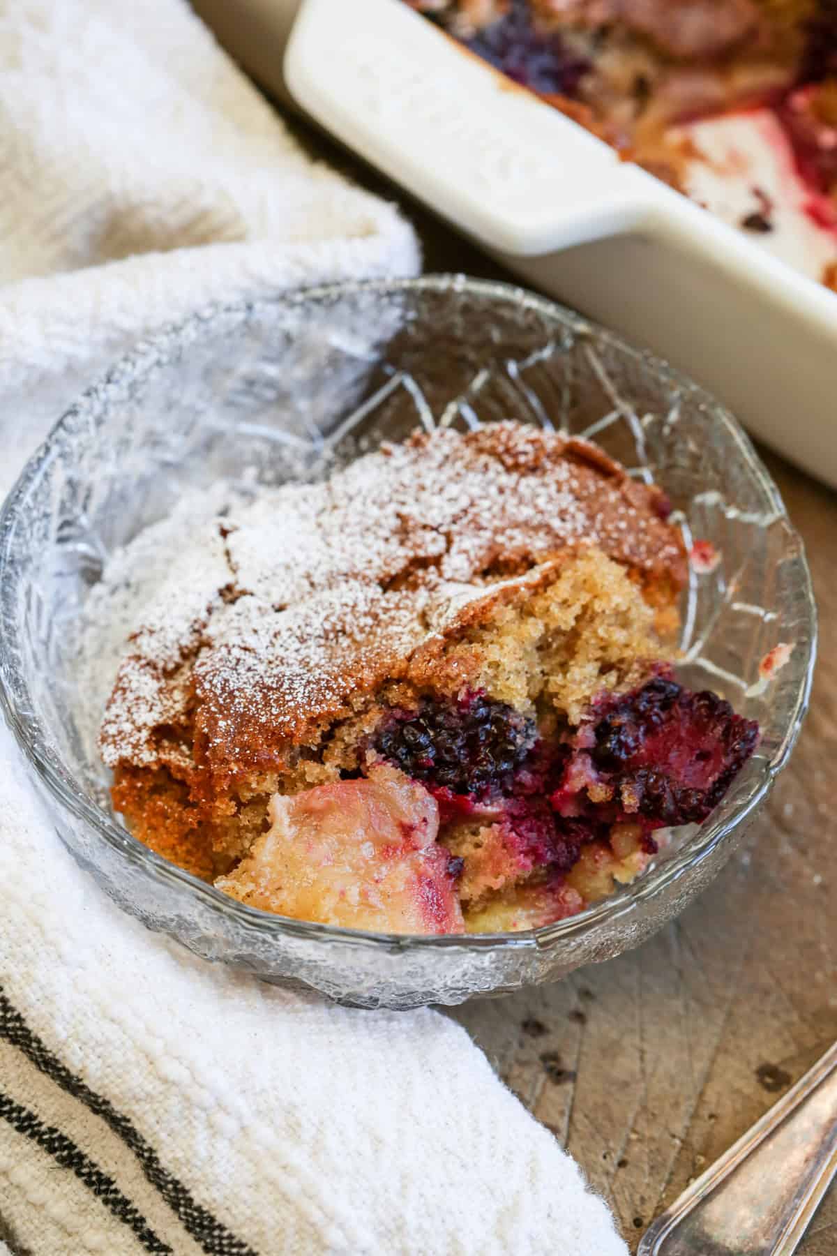Bowl of Gluten-Free Blackberry Apple Sponge Pudding showing icing sugar dredged top, soft golden sponge and a rich fruity layer of apples and blackberries.