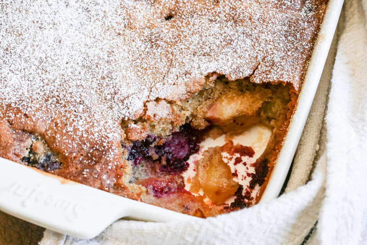 Serving dish of apple and blackberry pudding with large serving taken out showing golden sponge giving way to juicy fruity layer.