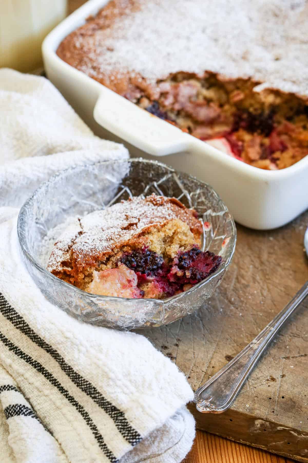 Bowl of Gluten-Free Blackberry Apple Sponge Pudding showing icing sugar dredged top, soft golden sponge and a rich fruity layer of apples and blackberries.