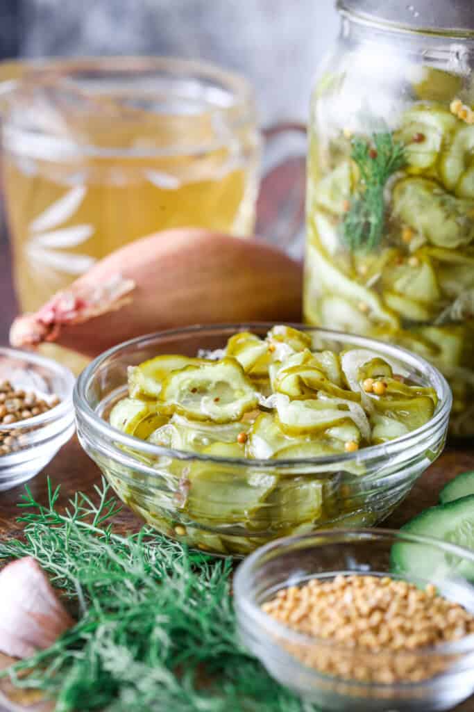 A small bowl of of pickled cucumber and shallot surrounded by the ingredients of the bread and butter pickles.