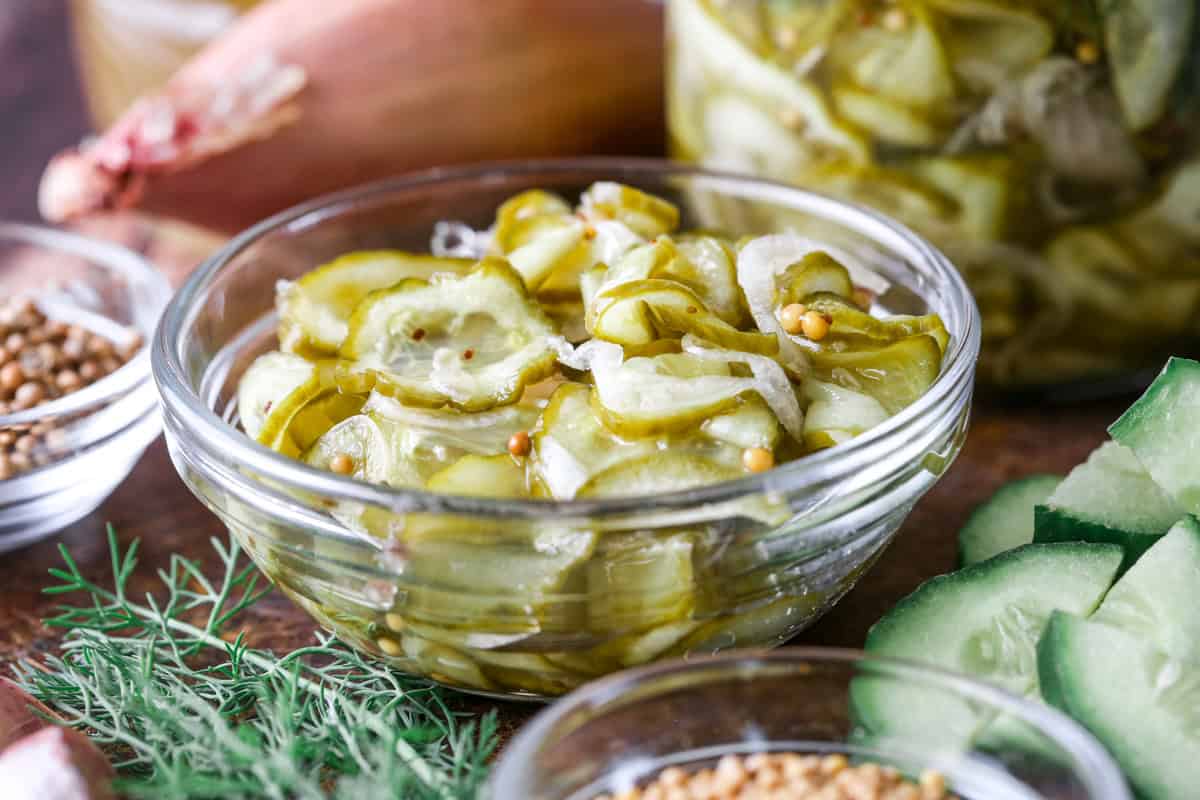 A small bowl of of pickled cucumber and shallot surrounded by the ingredients of the bread and butter pickles.
